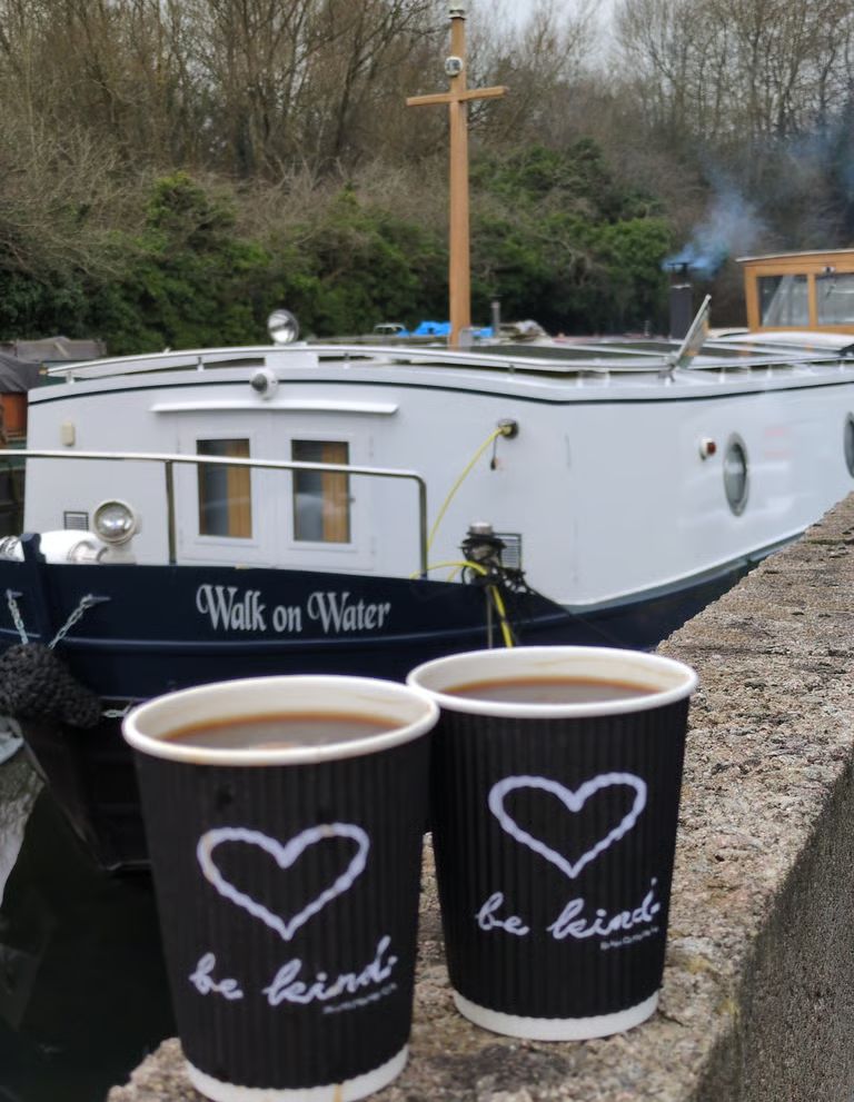The narrowboat Walk on Water moored on the canal, with a wooden cross at her mast, smoke from the chimney and two cups of tea on the wall
